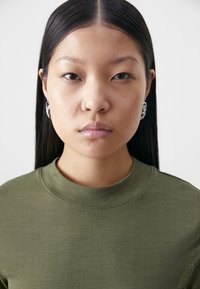 Chemise vert olive à col montant, en tissu texturé. Le modèle porte des boucles d'oreilles en argent avec des détails intricats.