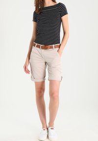 Shorts beige clair avec revers, associés à un haut rayé noir et blanc. Ceinture marron texturée et baskets blanches complètent la tenue.
