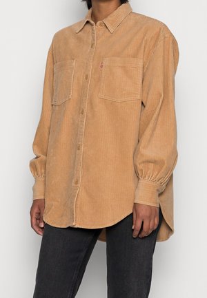 Personne portant une chemise en velours côtelé beige oversize à boutons avec deux poches poitrine et un pantalon noir, vue des épaules jusqu’aux hanches.