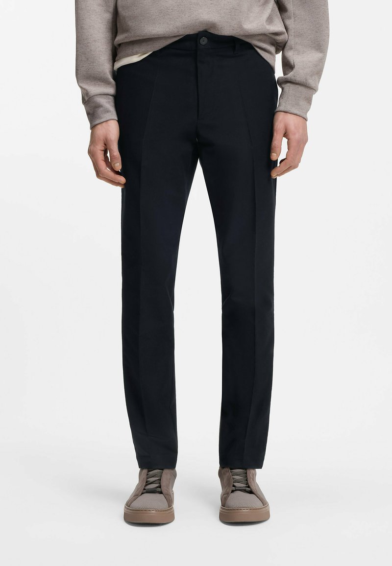 Μαύρο παντελόνι με εφαρμογή κοψίματος, με κουμπωτό κλείσιμο, πιέτες στο μπροστινό μέρος και ομαλή υφή υφάσματος, συνδυασμένο με γκρι αθλητικά παπούτσια.