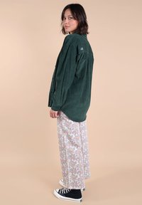 Chemise en velours côtelé vert avec des boutons à l'avant, associée à un pantalon fluide à motif floral et des baskets noires, sur fond neutre.