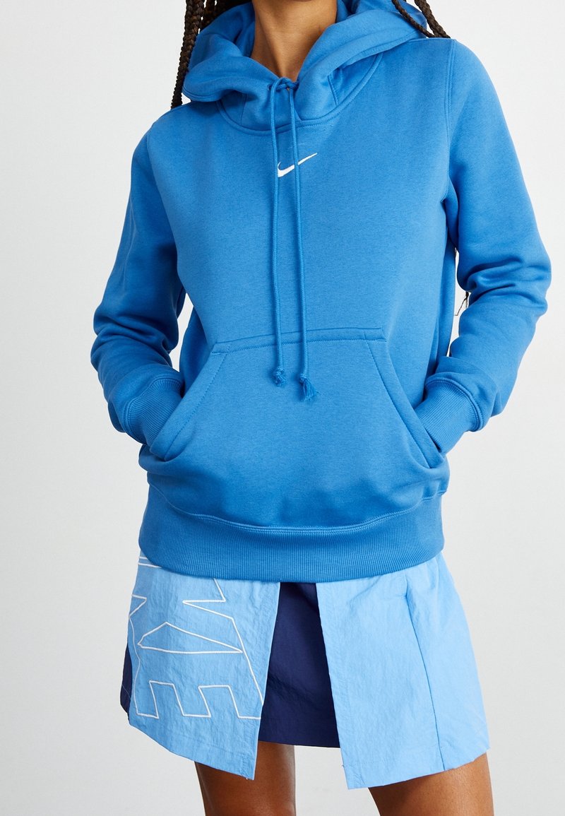 Blå Nike-hoodie med ficka fram, snörning i huvan och vit logotyp. Kombinerad med en lager-på-lager blå kjol med en vit grafisk design.