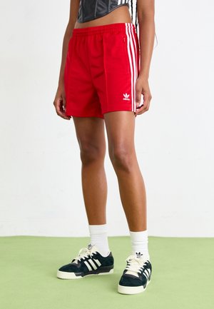 Person trägt rote Adidas-Shorts mit weißen Streifen, schwarze Adidas-Sneaker mit weißen Schnürsenkeln und weiße Adidas-Socken und steht auf einem grünen Boden.