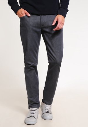 Mand iført mørkegrå slim-fit bukser, sort langærmet top og lysegrå sneakers, stående med den ene hånd i lommen.