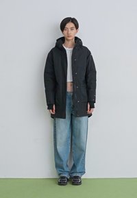 Jovem em pé, vestindo um parka preto com capuz, um top branco curto, jeans azuis largos e ténis pretos, contra um fundo simples.