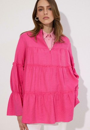 Γυναίκα που φοράει φαρδιά, λαμπερή ροζ μπλούζα με στρώσεις πάνω από ένα ανοιχτό ροζ πουκάμισο με γιακά, με ασημένια σκουλαρίκια κρίκους, στέκεται μπροστά σε απλό φόντο.