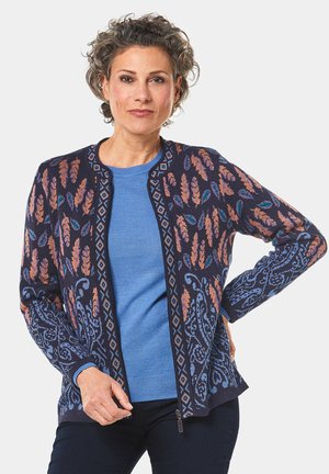 Cardigan bleu foncé avec des motifs floraux orange et bleu-vert, présentant un zip à l'avant, un col rond et des manches longues. Porté par-dessus un haut bleu clair.