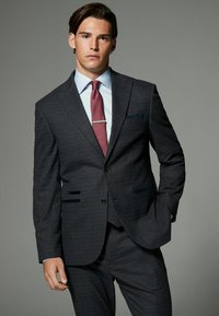 Costum gri închis cu carouri, cu închidere cu două butoane, rever cu tăietură și buzunare oblice. Asortat cu o cămașă de dress bleu deschis și o cravată burgundă.