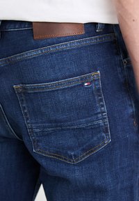 A kék farmernadrág slim fit szabással rendelkezik, sárga varrással és barna bőr címkével a hátsó részén. Egy kis logó látható a hátsó zseben.