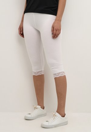 En person iført hvide korte leggings med blondekant og hvide sneakers, stående mod en ensartet lys baggrund.