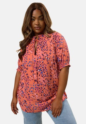 Vrouw draagt een oranje bloemenblouse met striksluiting en korte mouwen, gecombineerd met lichtblauwe spijkerbroek, staand tegen een witte achtergrond.