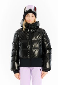 Zwarte pufferjack met een glanzende afwerking, voorrits, twee zijzakken en een aansluitende zwarte tailleband. Draagt een paar goggles en een gebreide beanie.