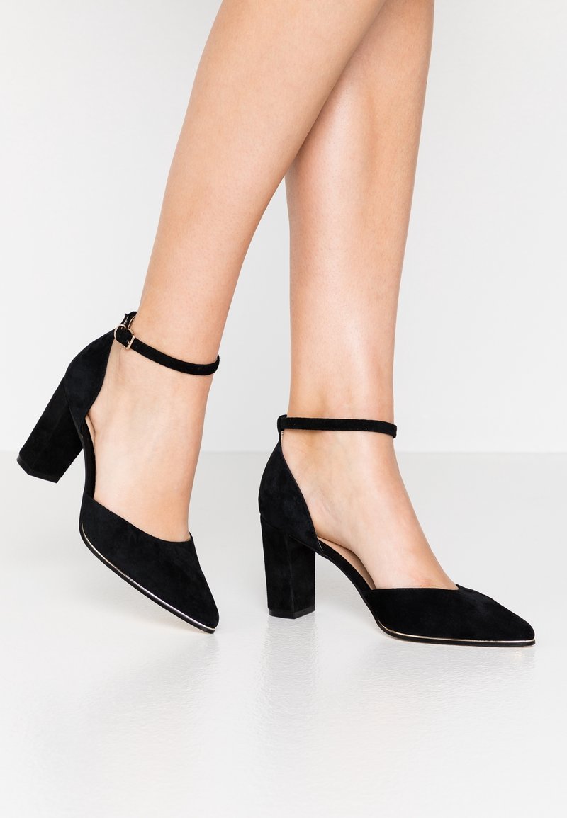 Chaussures à talons hauts en daim noir avec brides à la cheville portées aux pieds, montrant des bouts pointus et des talons blocs sur un sol blanc réfléchissant.