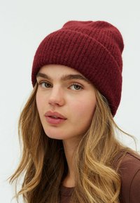 Căciulă tricotată în nuanță de vișiniu, cu o textură ribbed, dintr-un material moale și cu o ajustare ușor lejeră. Are un vârf rotund și o margine pliată.