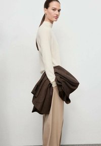 Beige Rollkragenpullover kombiniert mit weiten braunen Hosen. Ein dunkelbrauner Mantel hängt über dem Arm, hat eine glatte Textur und dezente Details.