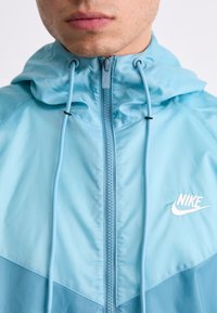 Gaiši zils Nike jaku ar kapuci, ar pilnu rāvējslēdzēju, regulējamām auklām un baltu logotipu uz krūšu daļas. Gluda, viegla auduma.