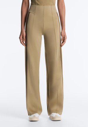 Pantalon beige taille haute à jambes larges avec coutures verticales, associé à des baskets blanches sur une personne debout devant un fond uni.