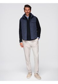 Gilet trapuntato navy sopra un maglione nero a trecce, camicia azzurro chiaro con colletto, pantaloni crema e sneakers beige.
