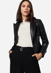 Schwarze Leder-Motorradjacke mit gerippten Schulterakzenten, Reißverschluss und langen Ärmeln, getragen über einem weißen Hemd und schwarzen Hosen.