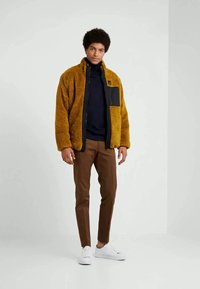 Young man wearing a mustard yellow fleece jacket over a black turtleneck, brown pants, and white sneakers, standing against a plain background.