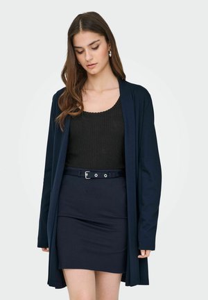 Kvinne iført en svart ribbestrikket topp, marineblå skjørt med belte, og matchende lang marineblå åpen cardigan, stående mot en ensfarget bakgrunn.