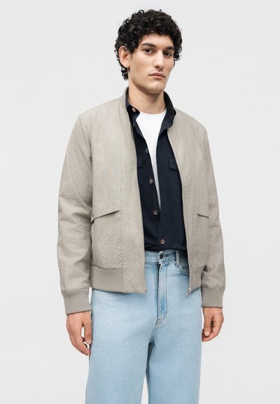 Homme aux cheveux bouclés portant une veste bomber beige texturée sur une chemise noire et un tee-shirt blanc, associé à un jean bleu clair, sur fond uni.