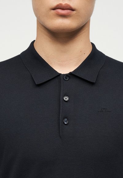 Gros plan sur une personne portant un polo noir à col avec trois boutons noirs et un logo brodé subtil sur la poitrine.