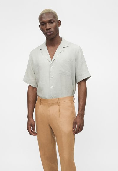 Άνδρας με κοντά ξανθά μαλλιά που φορά ανοιχτό γκρι πουκάμισο με κοντά μανίκια και μπεζ παντελόνι με ψηλή μέση σε λευκό φόντο.
