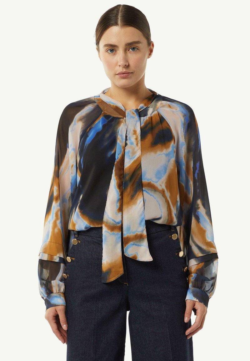 Bunt gemusterte Bluse mit blauen, braunen und cremefarbenen abstrakten Mustern. Lange Ärmel mit Manschetten; Bänder am Hals. Leichter, fließender Stoff.