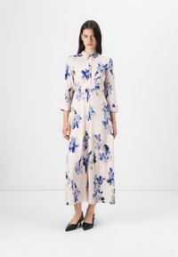 Modellen bär en blommig, pastellfärgad klänning med krage och knappar, i kombination med svarta klackar, stående mot en plain bakgrund.