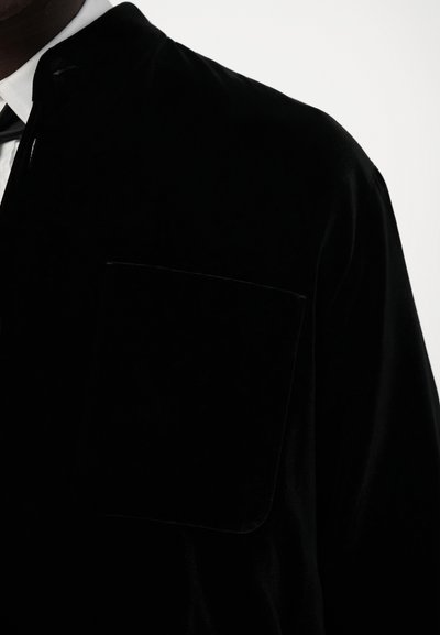 Black velvet shirt with a pointed collar, featuring a chest pocket. The texture is smooth and soft, emphasizing a sleek, modern design.