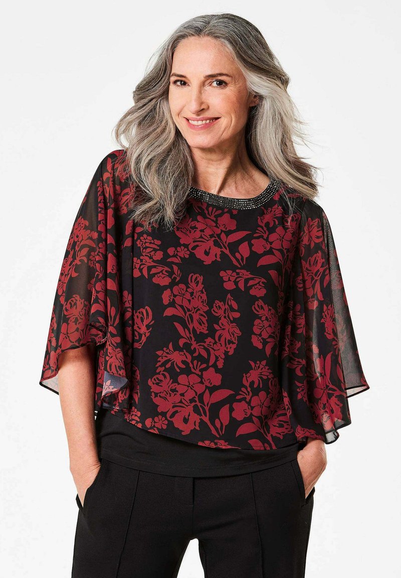 Blusa nera trasparente con motivo floreale rosso, maniche ampie e scollatura decorata con perline. Abbinata a pantaloni neri, che mostrano una vestibilità comoda.