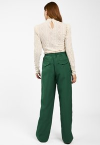 Groene wijde broek met een gladde textuur, voorzien van twee achterzakken. Het model draagt een crèmekleurige top met lange mouwen en een keyhole-detail.