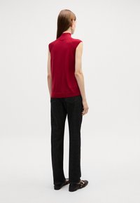 Top senza maniche rosso con colletto alto, abbinato a jeans neri a gamba dritta. Le calzature presentano dettagli con cinturini. Tessuto liscio e design semplice.