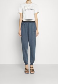 White t-shirt with "EA7 Emporio Armani" logo in gold, worn with blue joggers featuring elastic waist and ankle cuffs, paired with strappy sandals.