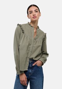 Chemise olive à boutons avec un col en volants et un détail de poitrail. Manches longues terminées par des poignets ajustés, faite dans un tissu texturé.