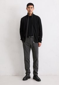 Schwarze Zip-Jacke über einem schwarzen Shirt, graue tapered Hose und schwarze Schnürstiefel vor einem einfarbigen Hintergrund. Strukturierte Stoffe sind sichtbar.