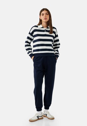 Langärmliges Polohemd in Marineblau und Weiß mit Streifen und Kragen, kombiniert mit Marineblauen Jogginghosen und weißen Sneakers mit schwarzen Akzenten.
