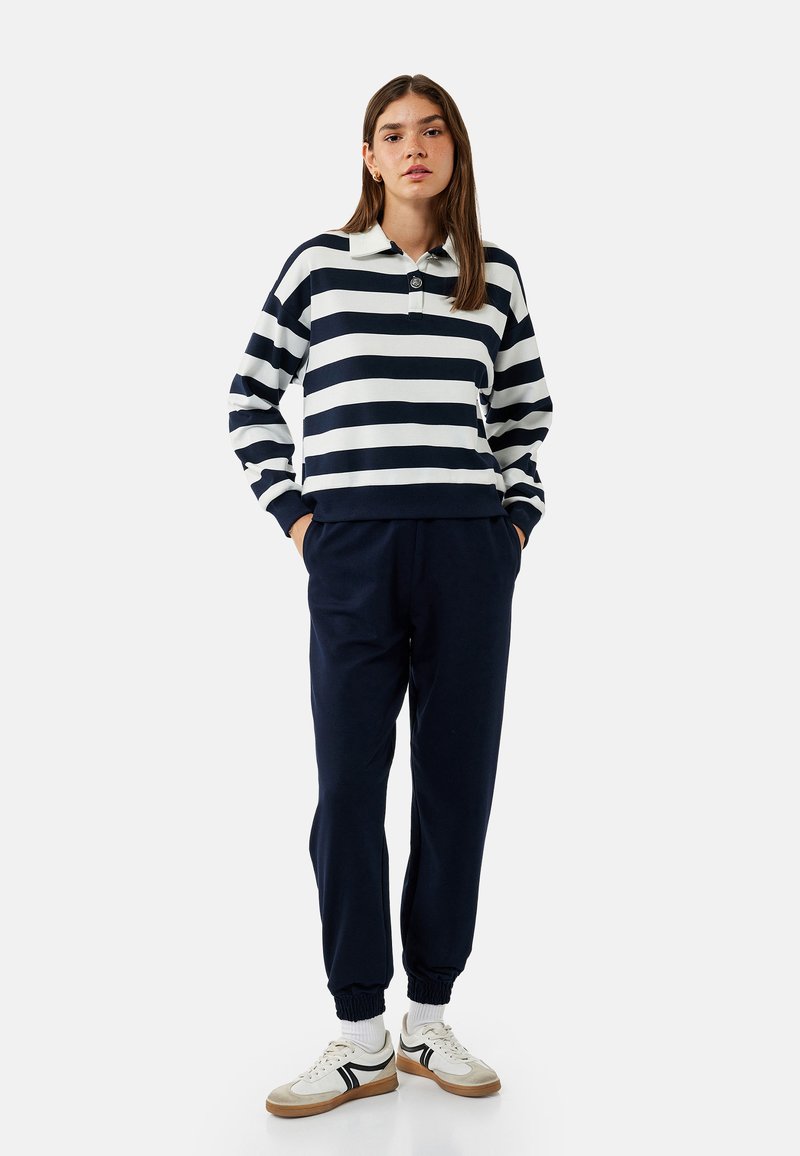 Langärmliges Polohemd in Marineblau und Weiß mit Streifen und Kragen, kombiniert mit Marineblauen Jogginghosen und weißen Sneakers mit schwarzen Akzenten.