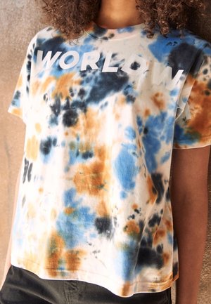 T-shirt tie-dye avec des taches bleues, orange et noires et le mot partiellement visible "WORLDW" sur la poitrine, porté par une personne.