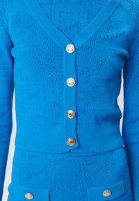 Cardigan lavorato a maglia blu con scollatura a V, dotato di bottoni dorati, motivo testurizzato e polsini a coste. Abbinato a una gonna coordinata.
