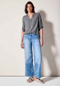 Blouse grise à manches longues avec une patte de boutonnage, associée à un jean bleu clair à jambes larges et des sandales à accents argentés. Ombre légère en arrière-plan.