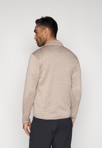 Μπεζ, υφασμάτινο πουλόβερ με ψηλό γιακά, μακριά μανίκια και ευθεία γραμμή στο τελείωμα, συνδυασμένο με σκούρο παντελόνι. Έμφαση στη ζεστασιά και την άνεση.