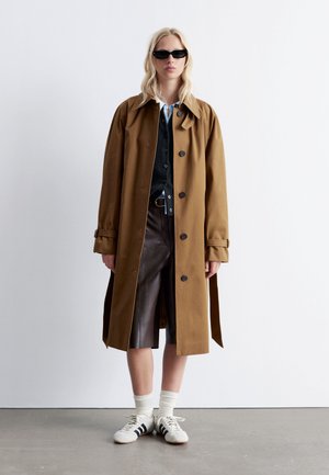 Brown trench coat with large collar and black buttons, layered over a black vest. Styled with leather shorts, white socks, and sneakers.