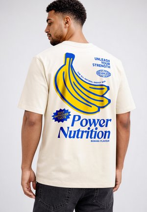 Man wearing beige T-shirt with large blue and yellow banana graphic and text promoting "Power Nutrition" energy bar, banana flavor.