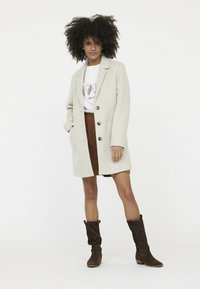 Beige Wollmischungs-Mantel mit taillierter Passform, drei Knöpfen und großen Taschen. Kombiniert mit einem braunen Cordrock und braunen lässigen Stiefeln.