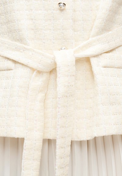 Ζώνη από ύφασμα με υφή κρέμας δεμένη σε χαλαρό κόμπο γύρω από ένα αντίστοιχο ενδύμα από τουίντ με πλισέ λευκό ύφασμα από κάτω.