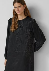 Schwarzes langärmliges Kleid mit einem strukturierten, wellenförmigen Muster. Es hat einen runden Ausschnitt und leicht aufgepuffte Ärmel für zusätzliche Details.