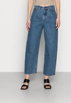 Personne portant un jean bleu taille haute et coupe ample avec un top court à pois et des sandales noires à bouts ouverts sur un fond uni.