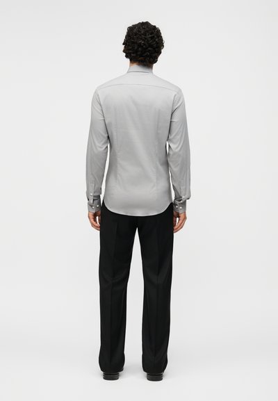 Άνδρας με σγουρά μαλλιά που φοράει ένα γκρι μακρυμάνικο πουκάμισο και μαύρο παντελόνι, στέκεται με την πλάτη προς το φακό σε μονόχρωμο φόντο.